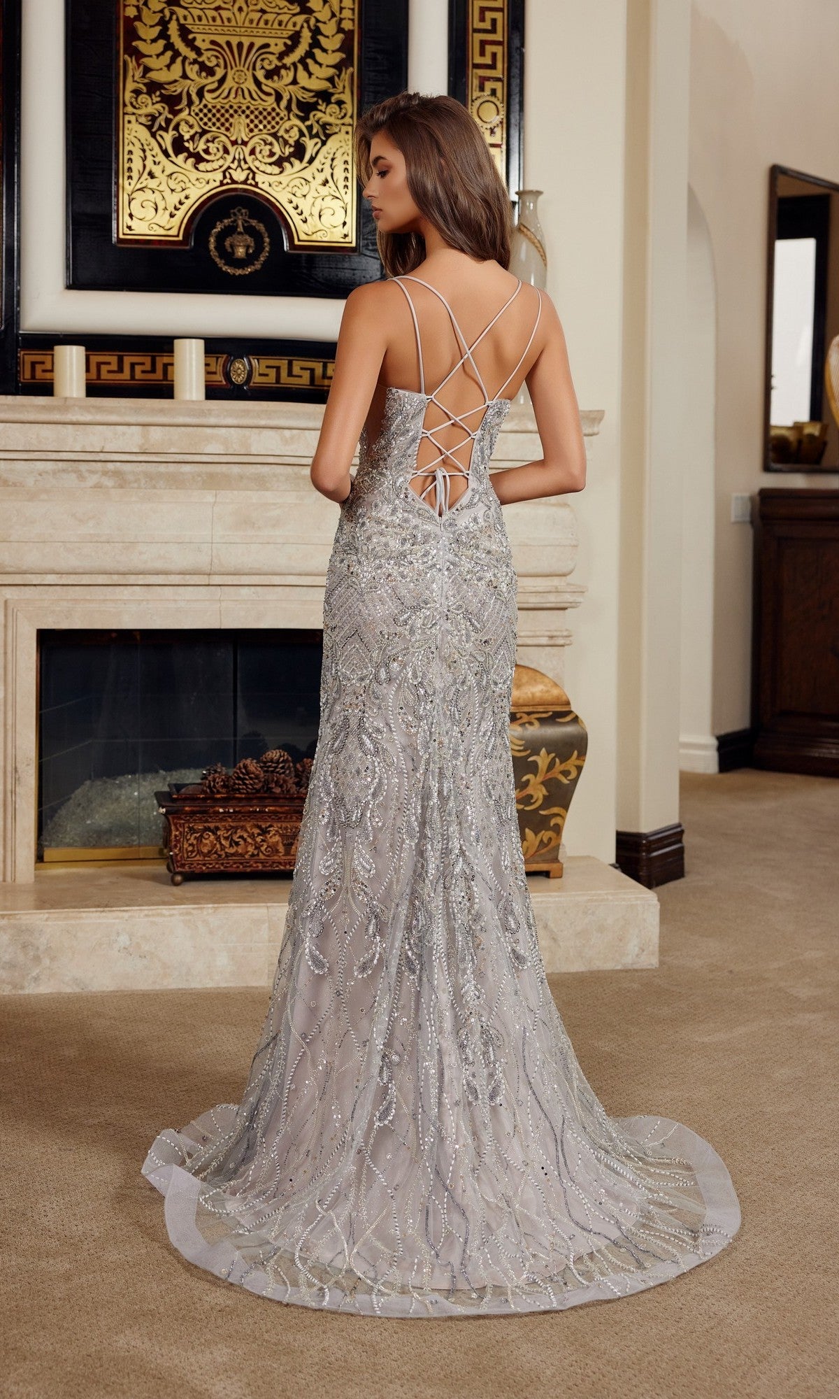 A woman stands indoors facing away from the camera, wearing the Nox Anabel C1834 long prom dress—a sleeveless sheath with intricate beadwork and crisscrossed straps.